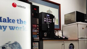Una máquina expendedora con el logotipo de Coca-Cola, un microondas y un refrigerador en una habitación con pared blanca y un cuadro enmarcado.