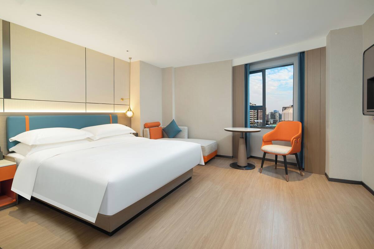 Ein Hotelzimmer mit einem großen Bett, einem kleinen Tisch, einem orangefarbenen Sessel und Blick auf die Stadt durch ein großes Fenster.