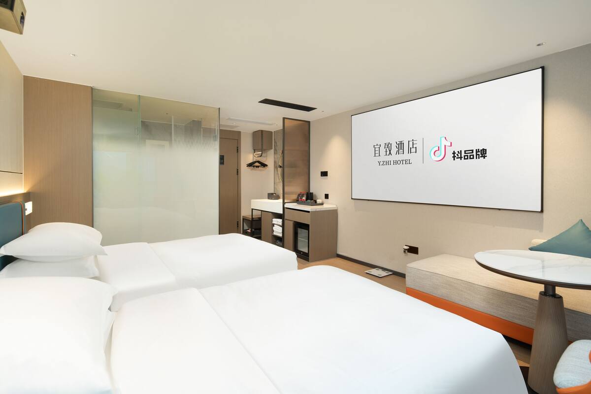 Ein modernes Hotelzimmer mit zwei Betten, einem kleinen Tisch und einem großen Bildschirm, der den Namen des Hotels zeigt.