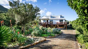 A two-story house with a balcony, surrounded by a garden with various plants and a paved pathway leading to the entrance. - House at Longbeach - upstairs apartment (Noordhoek, Chapmans Peak, Cape Town)