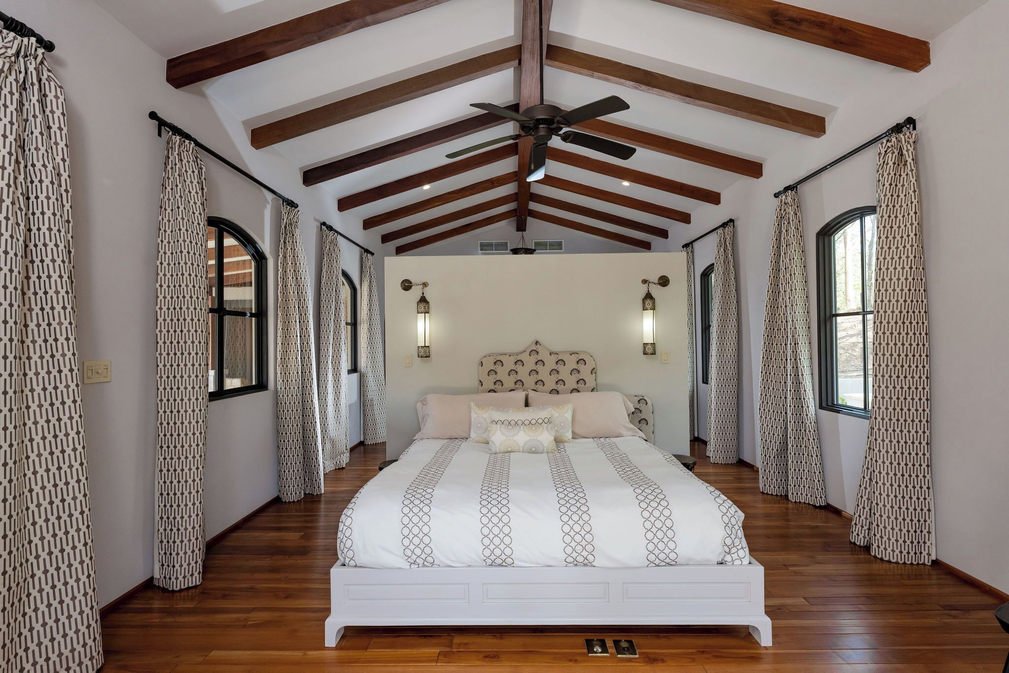 A bedroom with a large bed, wooden beams, patterned curtains, and wall sconces.