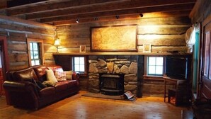 Interior de una cabaña de troncos con chimenea de piedra, sofá de cuero y suelos de madera.