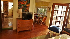 A room with a television on a wooden dresser, a dining table with chairs, a window with curtains, and a door with glass panels. - Lion's Gate Getaway- Lush Setting.  Check for seasonal discounts (St. Thomas,)