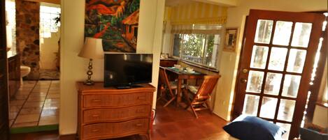 Quarto com televisão sobre um aparador de madeira, mesa de jantar com cadeiras, janela com cortinas e porta com painéis de vidro.