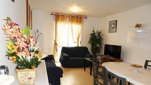 Ein Wohnzimmer mit schwarzem Sofa, einem Esstisch mit Stühlen, einem Fernseher und einer Topfpflanze.
