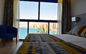 A hotel room with a large bed, a blue chair, and a view of a cityscape and beach. - Lido (Benidorm)