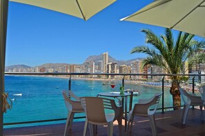 A balcony with a view of a beach and cityscape, featuring a table with chairs and a drink.
