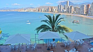 Une terrasse en bord de mer, avec des tables et des parasols blancs, offrant une vue sur une plage et le profil d’une ville, avec des montagnes en arrière-plan.
