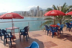 A terrace with outdoor seating, red umbrellas, and a view of a city skyline and beach.