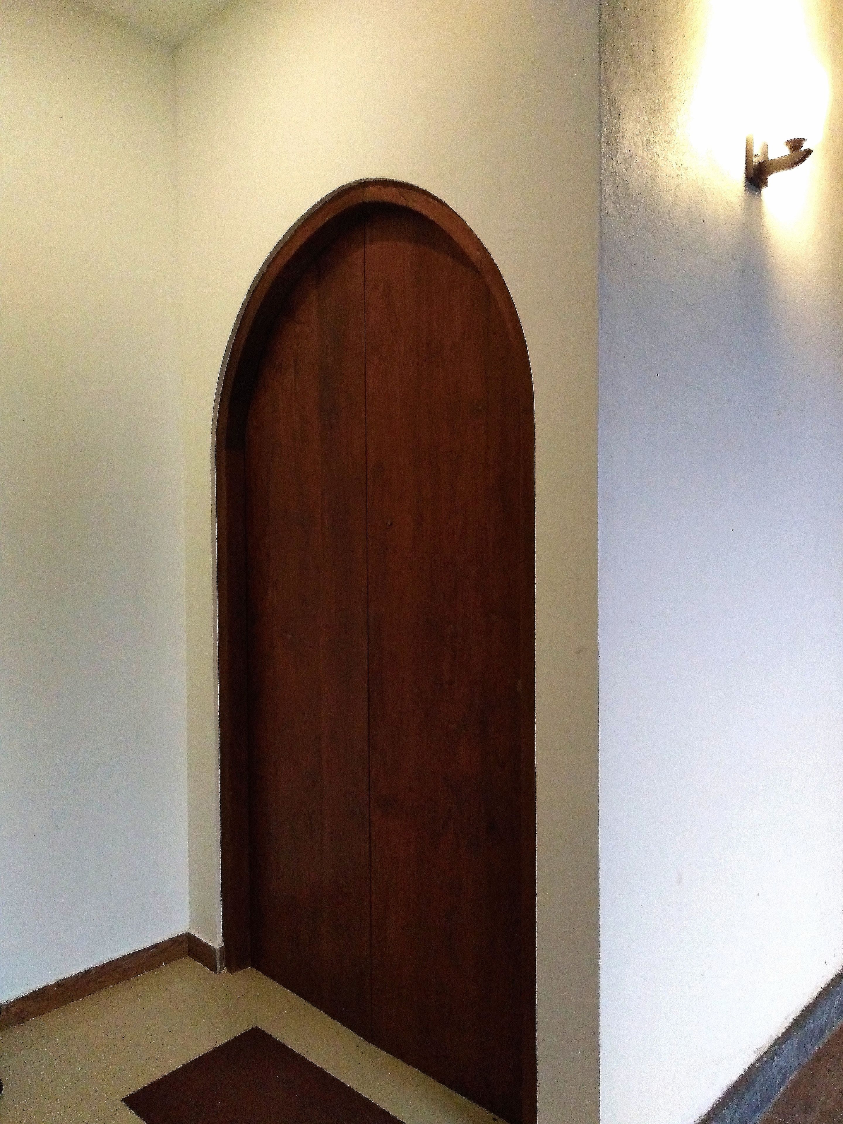 A closed wooden door with a simple arched top, a light switch on the wall, and a small rug at the door's base.