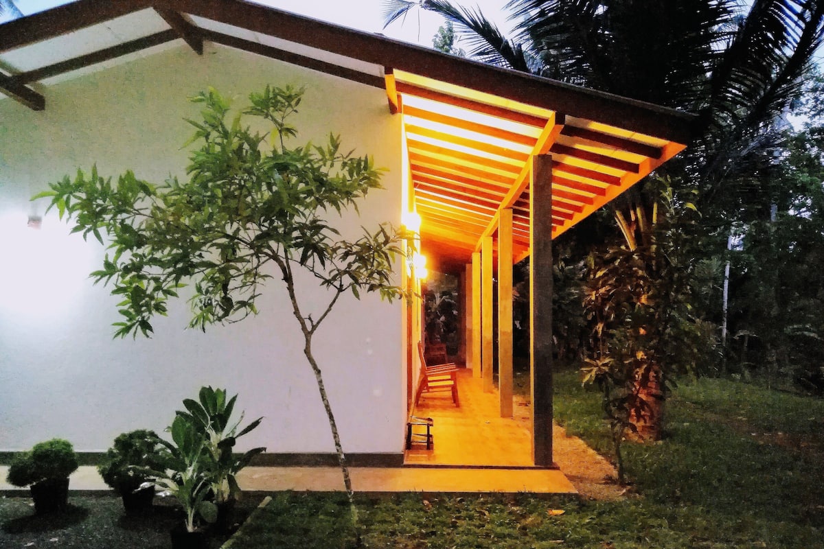 A modern building with a covered outdoor area, a pathway leading to a bench, and surrounding greenery.