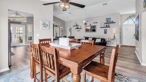 Un vaste espace à manger avec une table et des chaises en bois, un ventilateur de plafond et une vue sur le salon attenant.