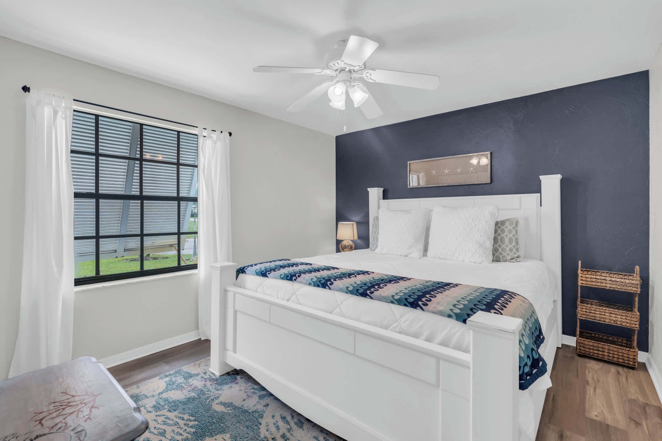 A bedroom with a bed, a ceiling fan, a window with curtains, a nightstand, and a wall-mounted shelf.