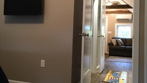 A hallway with a wooden floor, a flat-screen TV mounted on the wall, and a door leading to a living area with a couch and a window. - The Nest ~ Quiet location close to shops, dining and Downtown Freeport. (Freeport)