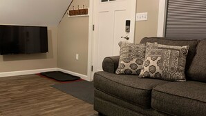 A living room with a gray sofa, patterned throw pillows, a wooden floor, a patterned rug, a flat-screen TV, a door, and an air conditioning unit. - The Nest ~ Quiet location close to shops, dining and Downtown Freeport. (Freeport)