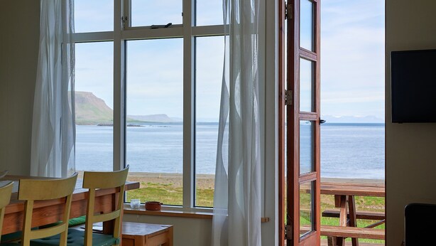 A room with a large window overlooking a beach and ocean, wooden furniture, and a view of a cliff.