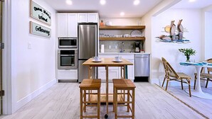Eine kleine Küche mit weißen Schränken, einem Edelstahlkühlschrank, einer Mikrowelle und einem Backofen. Es gibt einen Holztisch mit zwei Stühlen sowie einen kleinen Essbereich mit Korbstühlen.