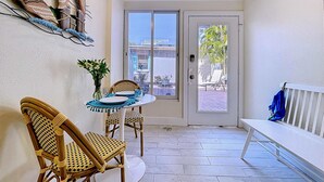 Ein Balkon mit Korbstühlen, einem Tisch mit Tellern und einer Blumenvase, mit Blick auf Gebäude und einen Baum durch die Tür.