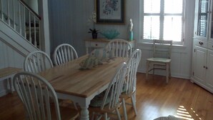 Ein Esszimmer mit Holztisch und weißen Stühlen, einer Treppe, einem Fenster mit Jalousien und einem Schrank mit Glastüren.