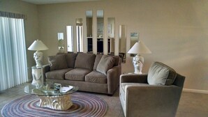 A living room with a brown sofa, a glass coffee table, and two lamps. There are mirrors on the wall and a patterned rug on the floor.