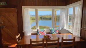Una mesa de comedor de madera con sillas, un cuenco de fruta y vistas a un lago a través de amplias ventanas.