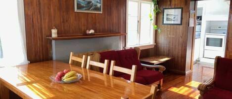 A wooden dining table with chairs, a red sofa, and a kitchen area with a refrigerator and shelves.