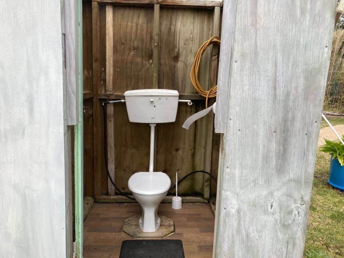 A small, enclosed wooden structure with a toilet, a hose, and a broom.
