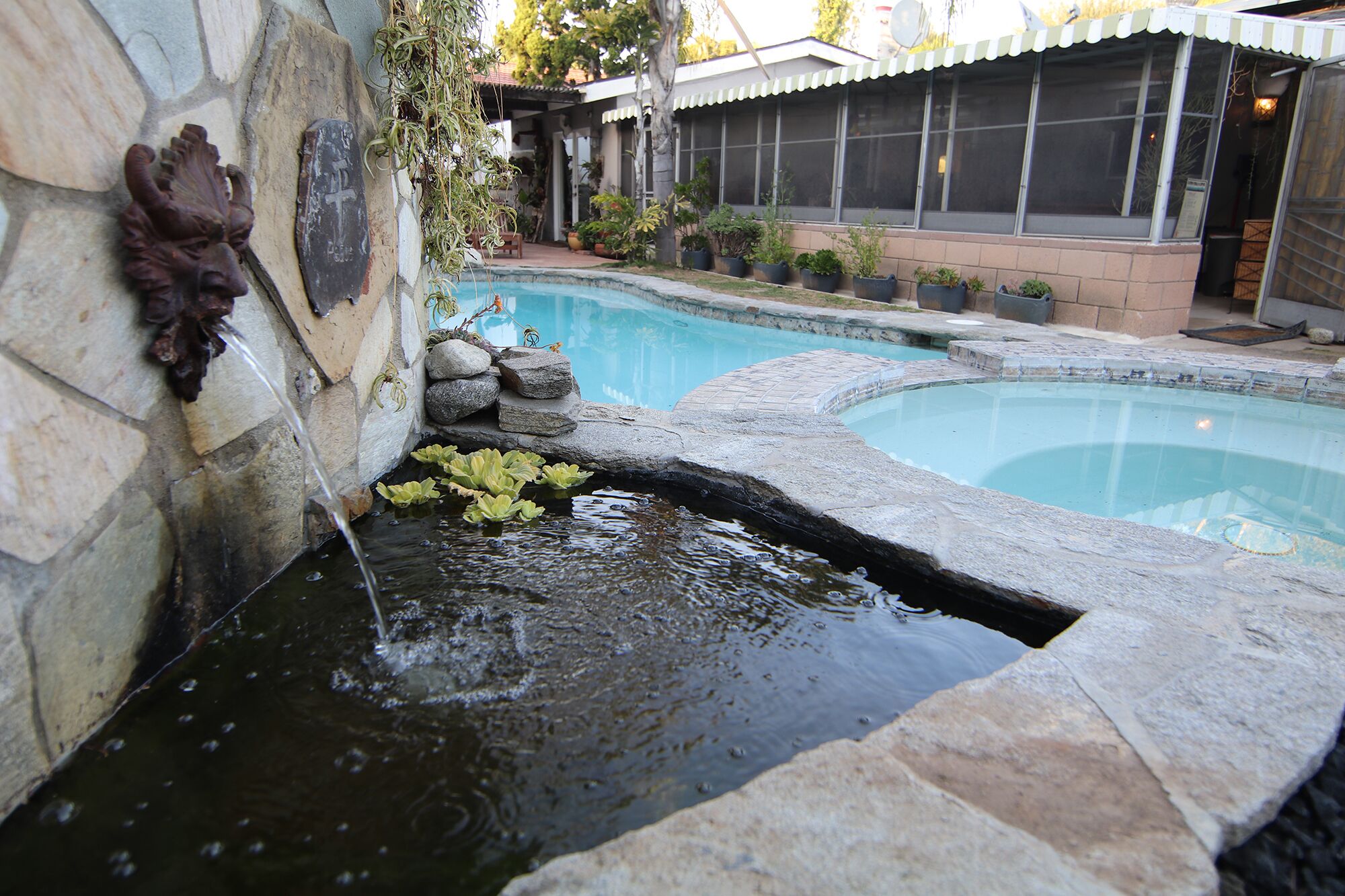 A swimming pool with a small waterfall feature, surrounded by stone walls and potted plants.