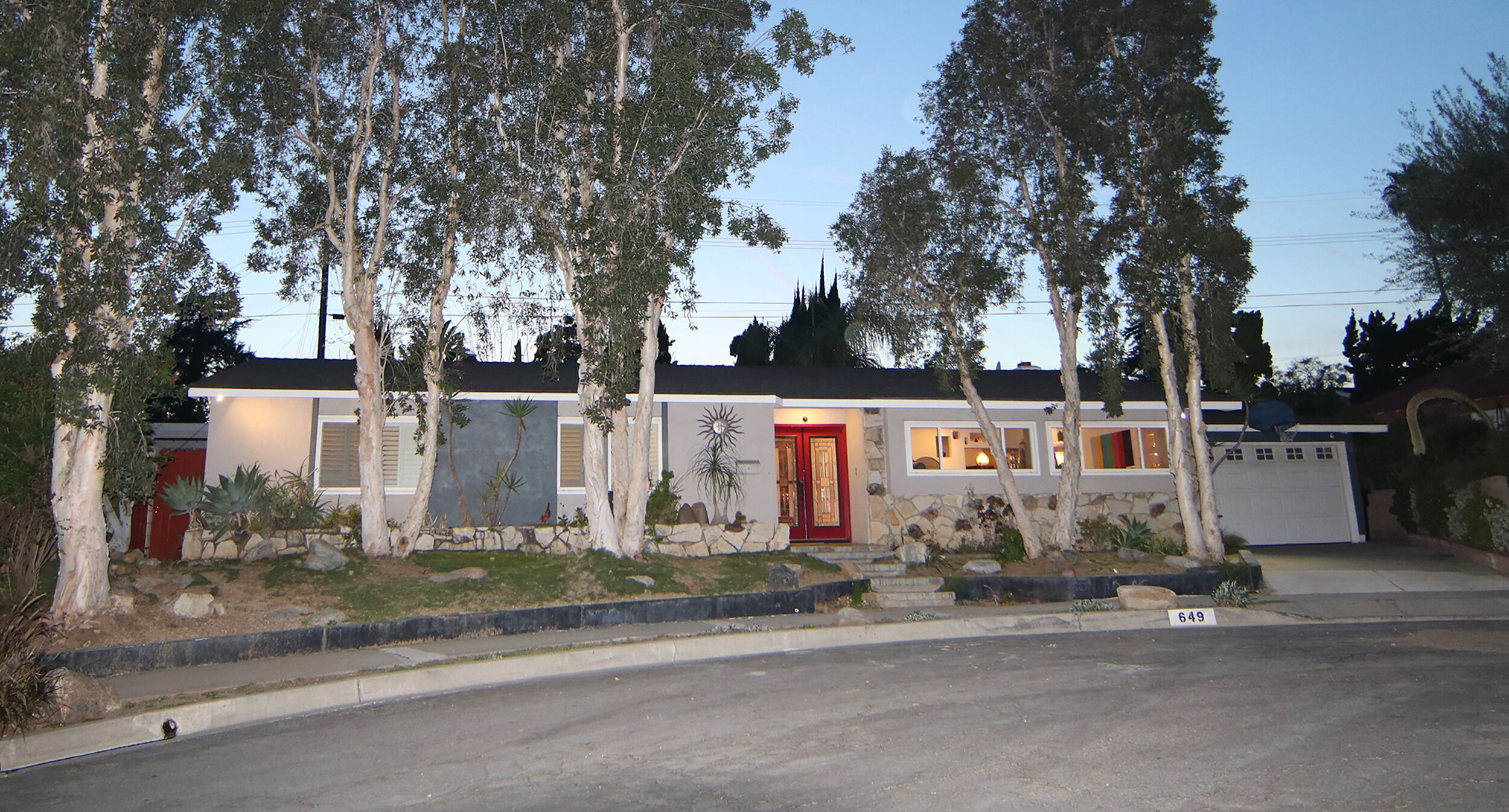 Una casa de dos pisos con puerta roja, un garaje y un jardín bien cuidado con diversas plantas y árboles.