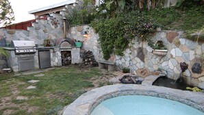 A backyard with a stone wall, a barbecue grill, a stone oven, a small pool, and a waterfall feature.