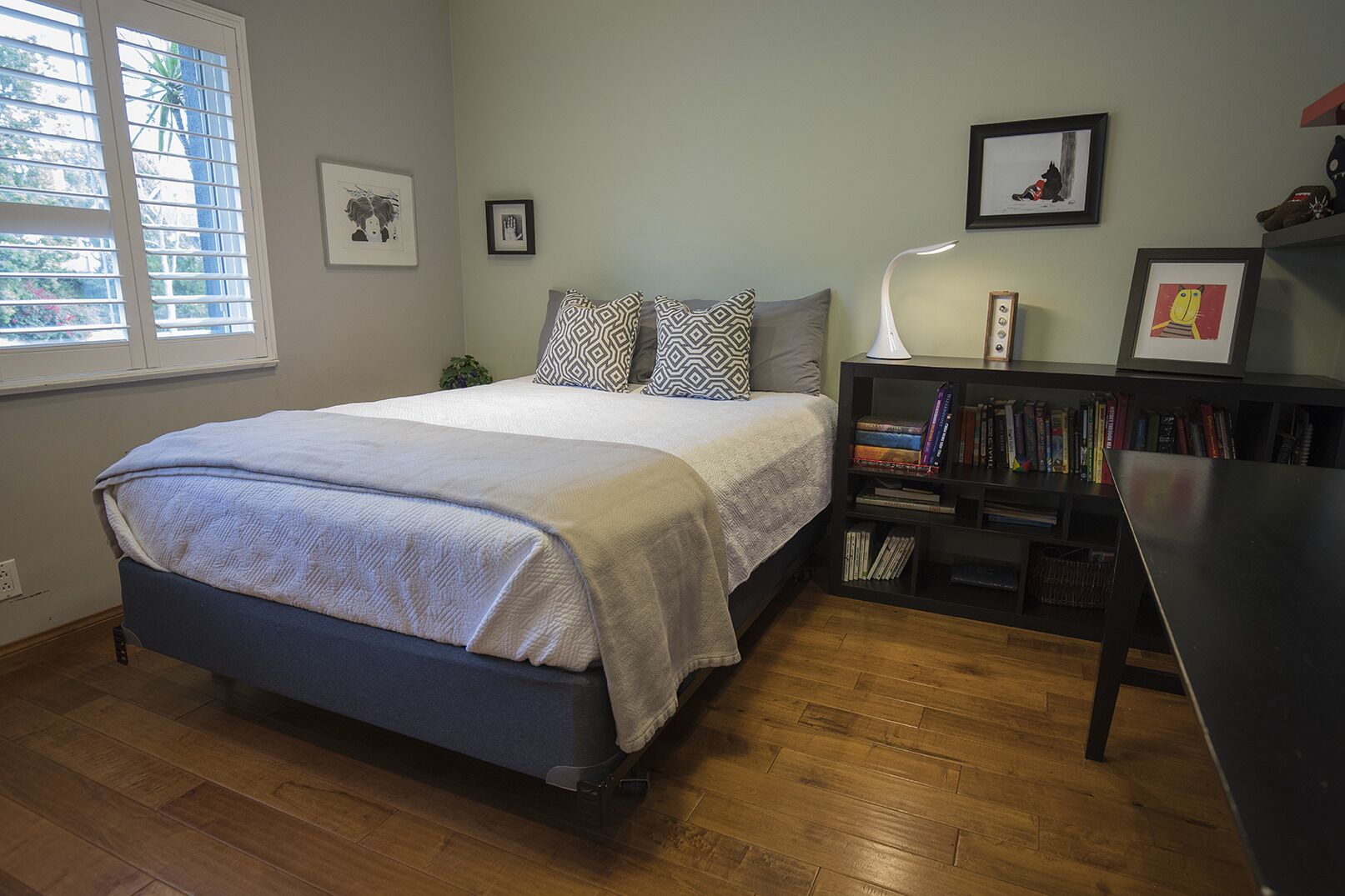 A bedroom with a bed, a bedside table with a lamp, a bookshelf, and a window with blinds.