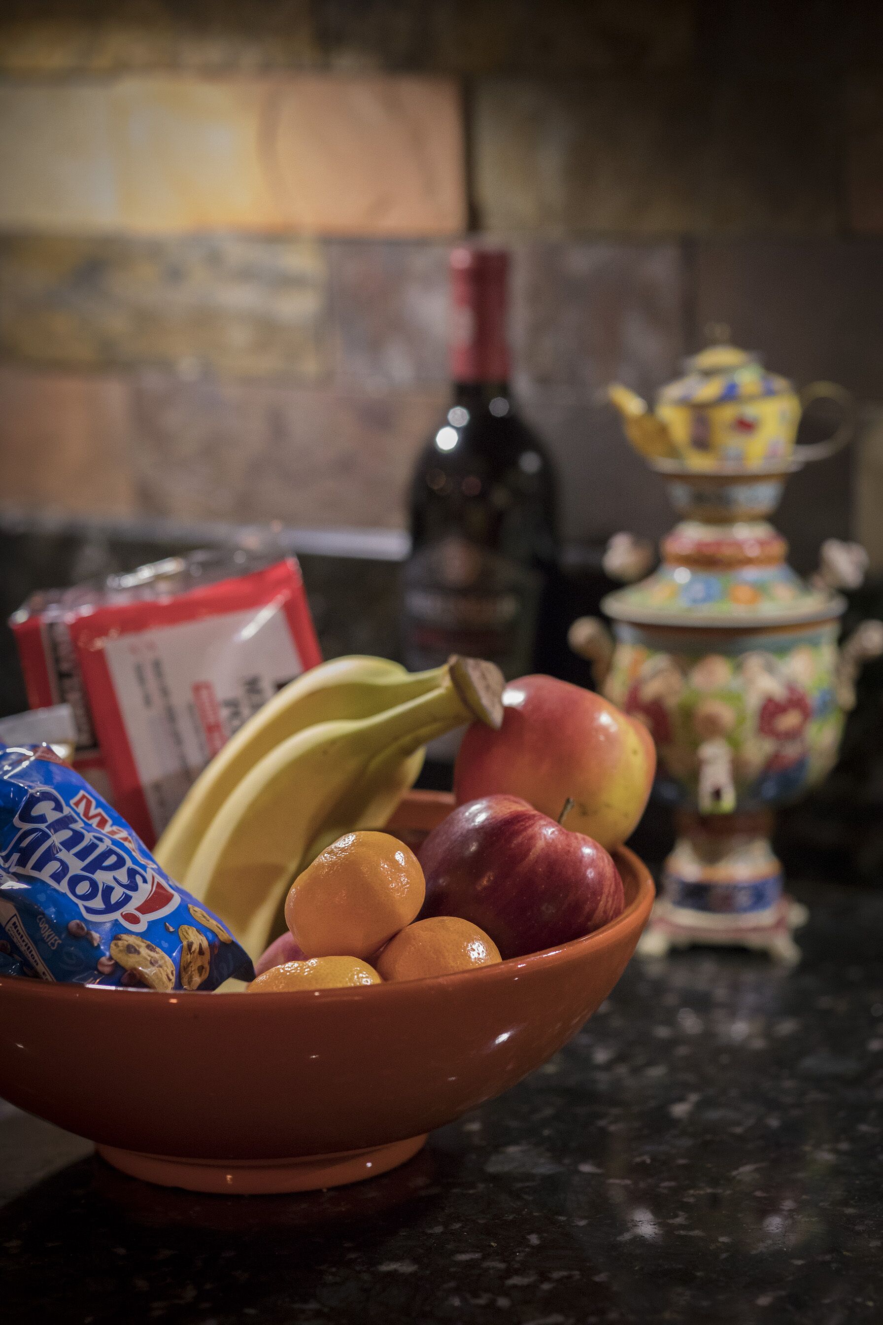 Uma tigela com frutas e petiscos em uma bancada da cozinha, com uma garrafa de vinho e um bule decorativo ao fundo.