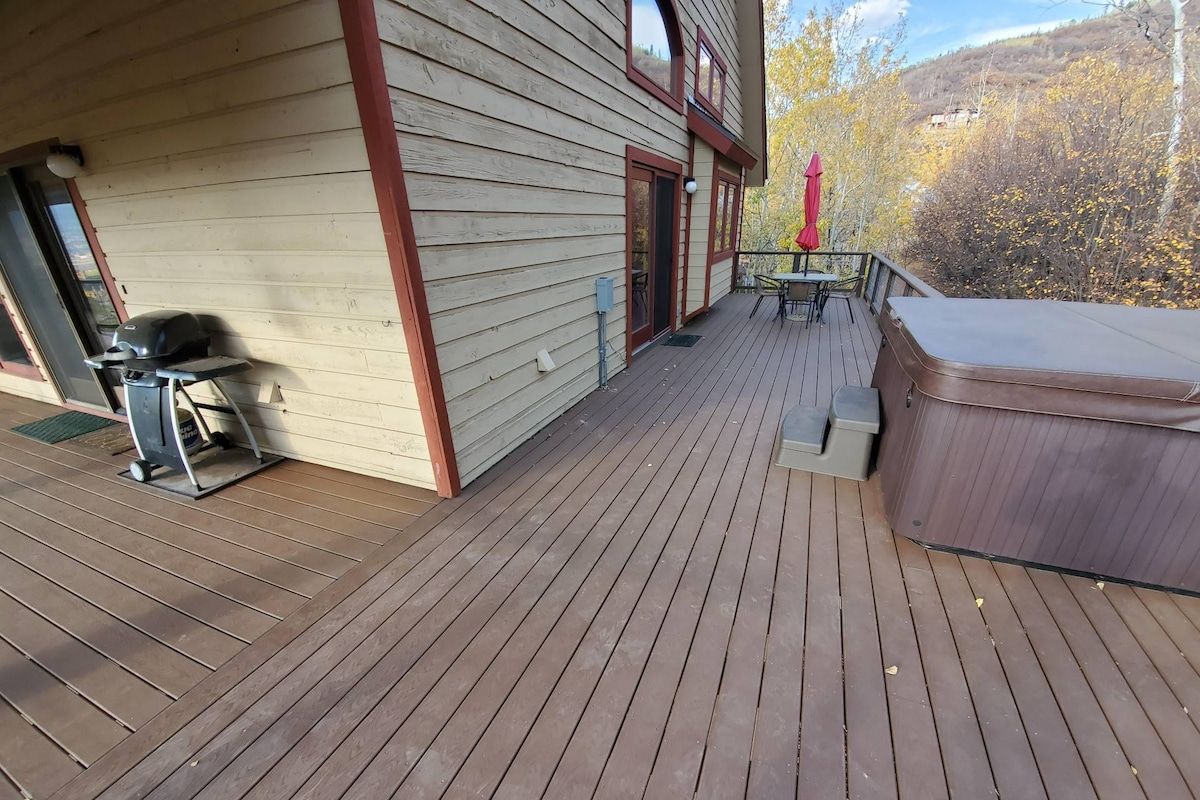 A wooden deck with a hot tub, outdoor seating, and a view of trees.