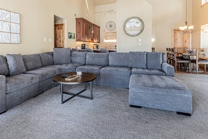A spacious living room with a large gray sectional sofa, a coffee table, and a dining area with wooden furniture.