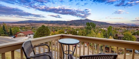 A balcony with wicker chairs and a small round table, overlooking a scenic landscape with mountains and a town.