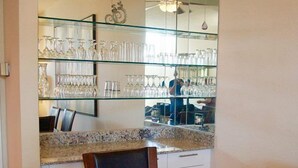 Una cocina moderna con una mesa de comedor de cristal, una zona de bar con estanterías llenas de copas, y una persona visible al fondo.