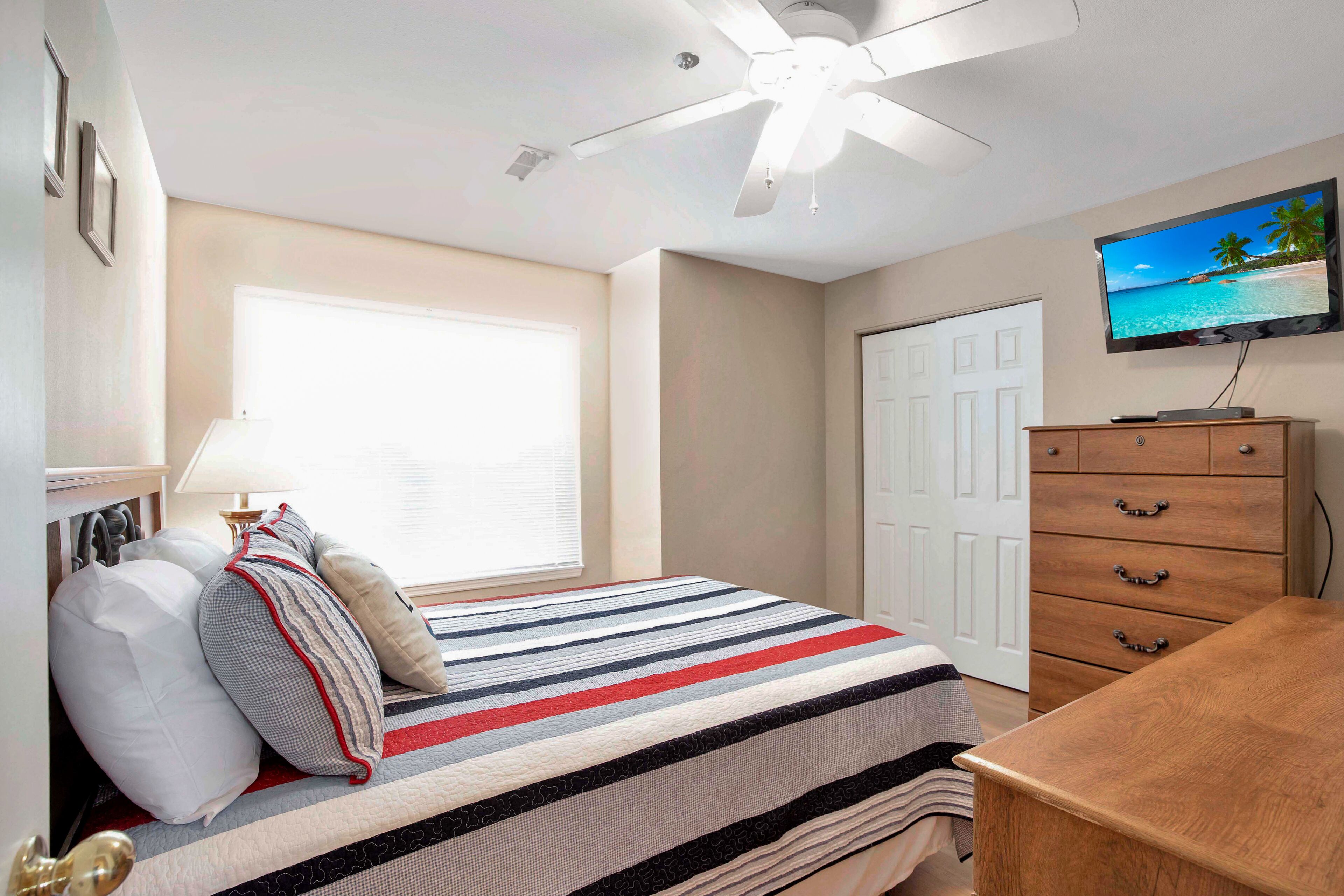 Een slaapkamer met een bed, een plafondventilator, een televisie aan de muur, een dressoir en een raam met jaloezieën.