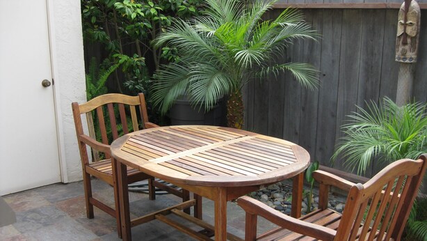 Una mesa redonda de madera con cuatro sillas, ubicada en un patio de piedra con una cerca de madera y vegetación al fondo.