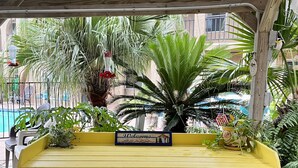 A yellow bench with a 'Welcome' sign under a wooden pergola, surrounded by lush greenery and a pool in the background. - 2 BR 2 bath -  pool - 3 minute walk to beach! (Corpus Christi)