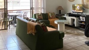 Sala de estar con un sofá verde, una mesa de centro y un televisor. Una puerta corrediza de vidrio da a un espacio exterior con sillas.