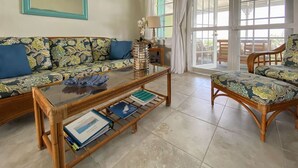Un salon comprenant un canapé, un fauteuil et une table basse en verre sur laquelle sont posés des livres. Une porte donne accès à un espace extérieur.