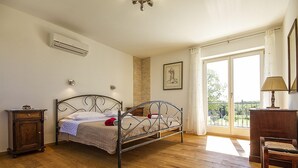 Un dormitorio con estructura de cama de metal, suelo de madera, una mesita de noche pequeña, una silla, un escritorio y un ventanal con vistas.