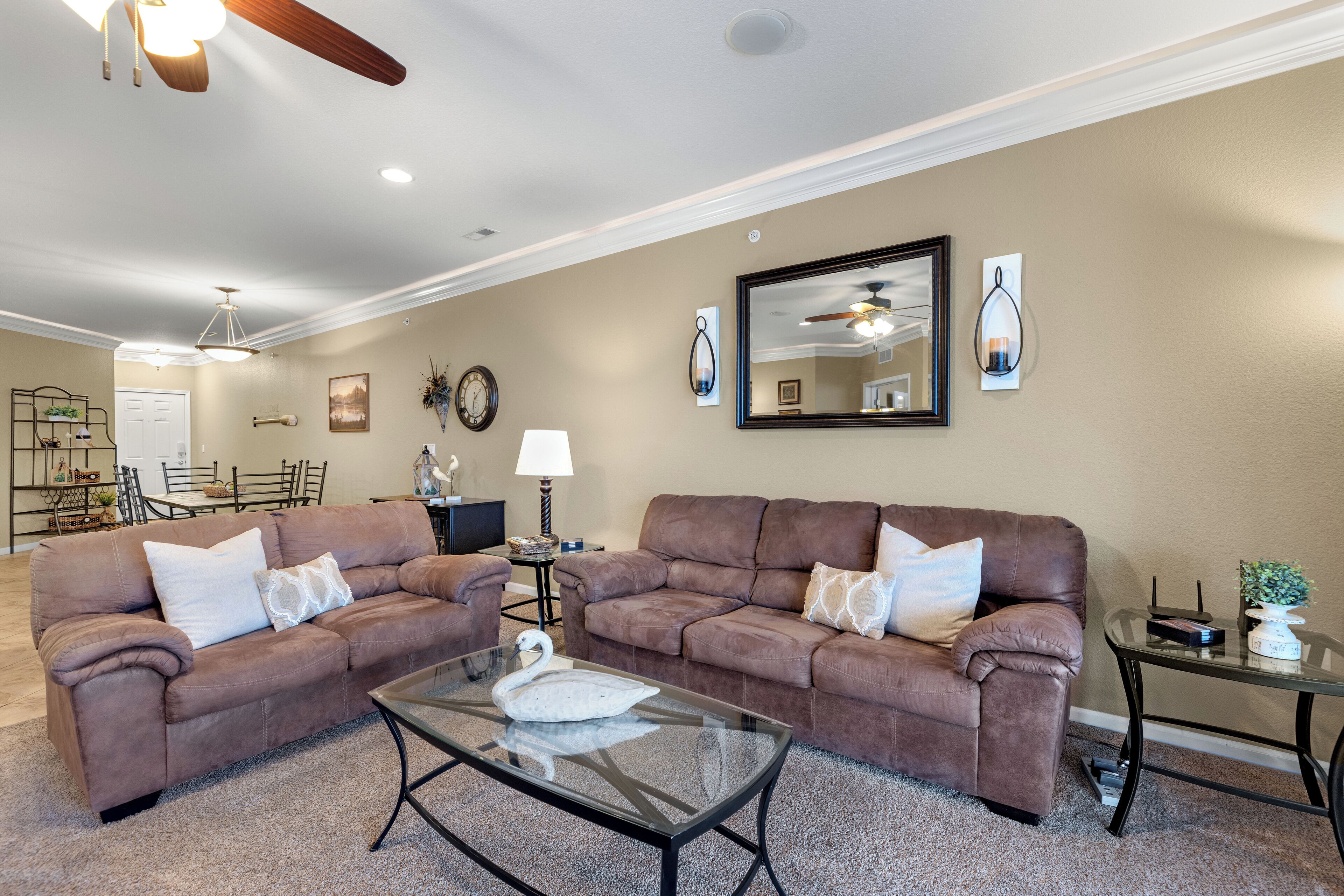A living room with two brown sofas, a glass coffee table, a ceiling fan, and a dining area in the background.