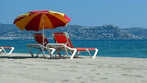 A beach with two red and white lounge chairs under a red and yellow umbrella, with a view of a coastal town and hills in the background. - 0086-BLAUCEL Apartment with seaview (Empuriabrava)