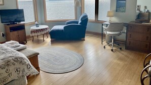 Een kamer met een groot raam met uitzicht op een wateroppervlak, een blauwe fauteuil, een tafeltje met een lamp, een televisie op een houten kast, en een bed met bloemenbeddengoed.