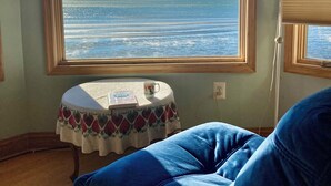 Een kamer met een groot raam dat uitzicht biedt op zee en een verre kustlijn. Er staat een klein tafeltje met een decoratieve tafelkleed en een kop erop. Een blauwe gestoffeerde stoel is op de voorgrond zichtbaar.