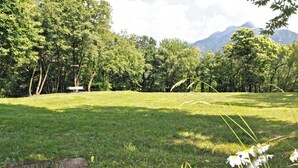 Un campo cubierto de hierba con un banco y un cartel al frente, rodeado de árboles y montañas en la distancia.