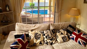 A room with a large window overlooking a swimming pool, a sofa with patterned cushions, and a shelf with decorative items.