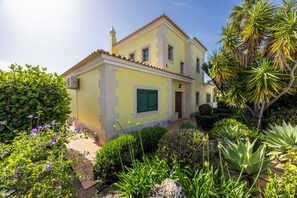 A two-story yellow building with a tiled roof, a small tower, and a garden with various plants and flowers.