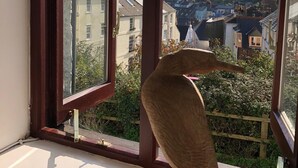 Een raam met uitzicht op een stad, een houten beeldje op de vensterbank, en een heldere lucht.